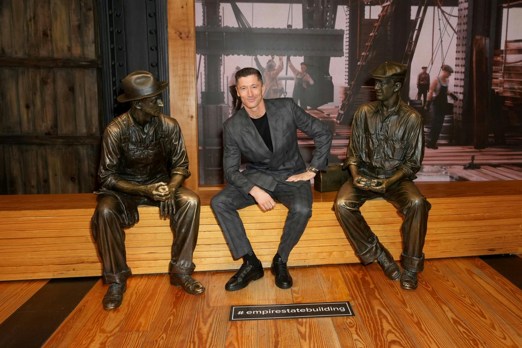 Muž v šedém obleku sedí mezi dvěma bronzovými sochami dělníků na dřevěné lavici. Cedule s nápisem „#empirestatebuilding“.