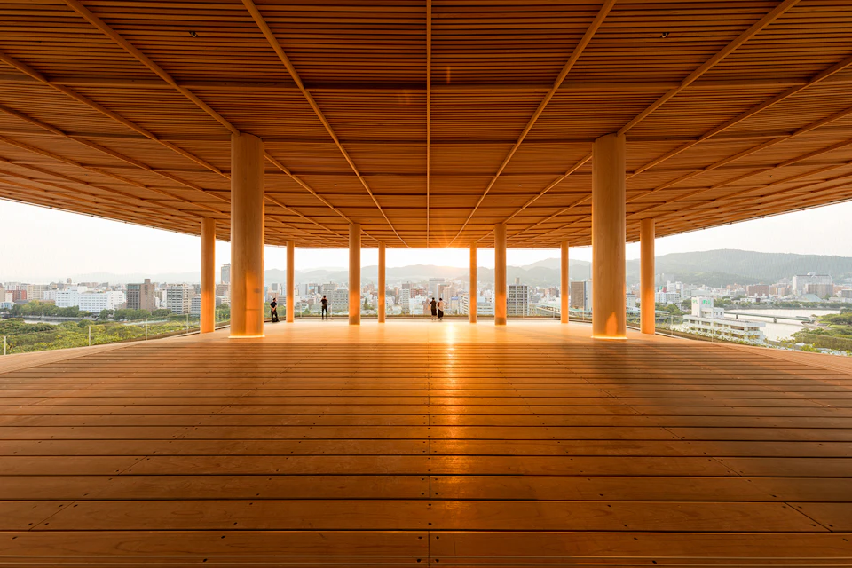 A large wooden pavilion with columns, overlooking a cityscape, with people in the distance at sunset.