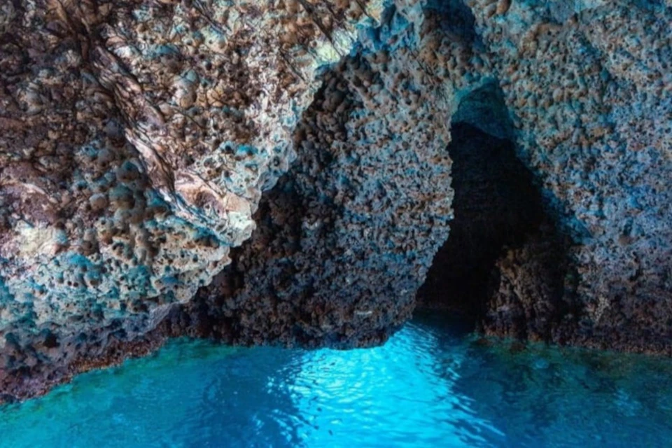 A rocky cave with turquoise water reflecting light, showing textured rock walls and an opening leading to darkness.