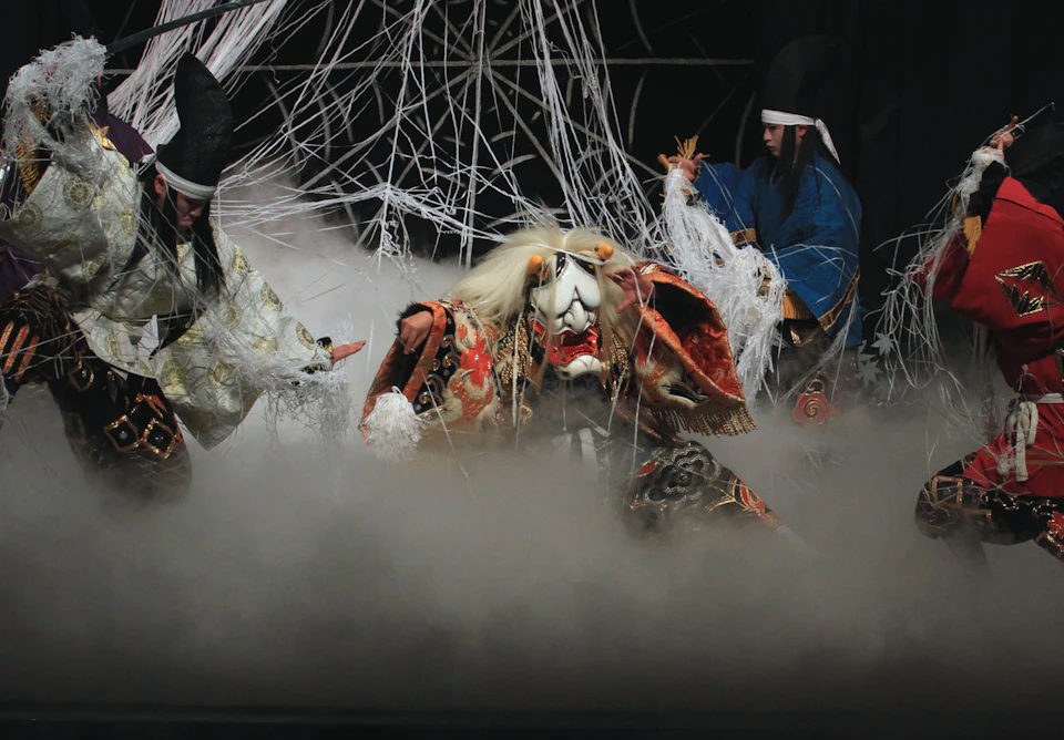 Performers in elaborate costumes on stage with dramatic lighting, fog effects, and white string decorations.