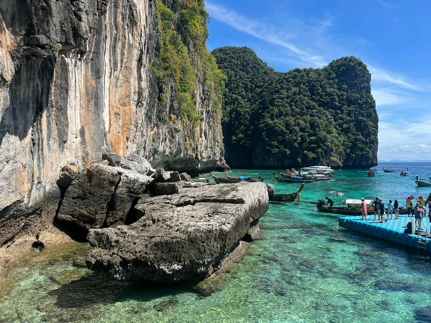 Une eau claire et turquoise, des bateaux amarrés près d'un rivage rocheux avec des falaises, des collines verdoyantes en arrière-plan et un ciel d'un bleu éclatant.