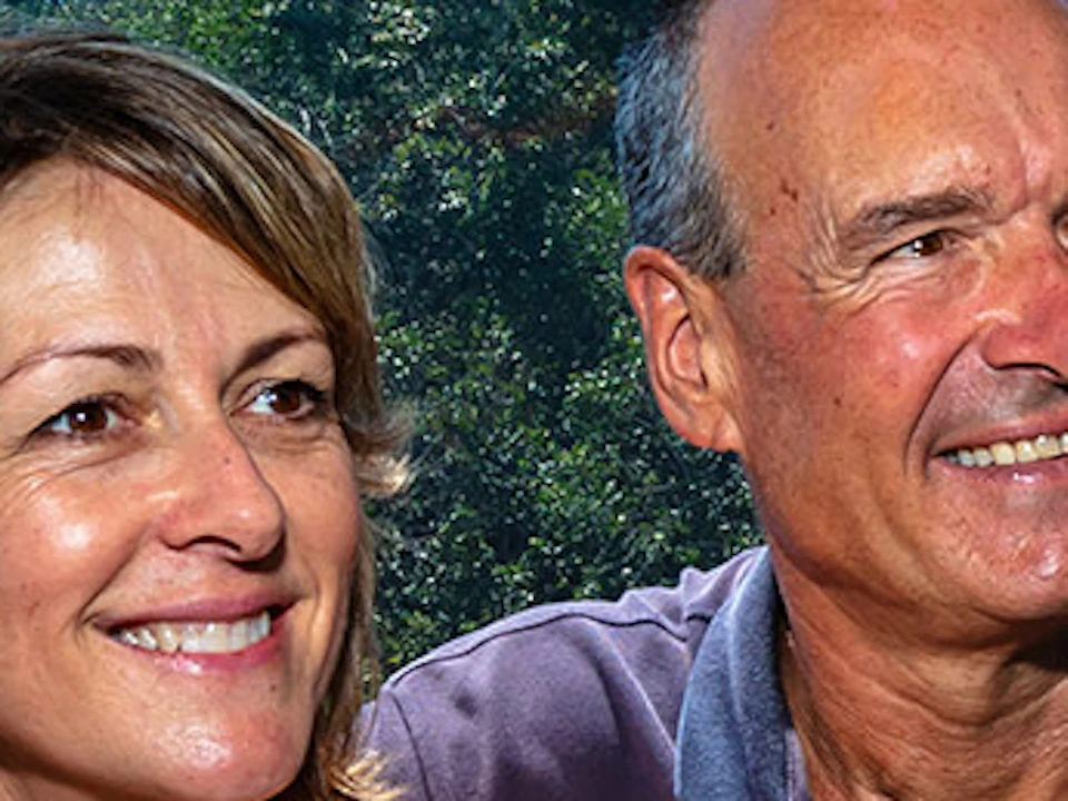 A smiling man and woman sit in a green cable car with a scenic view of lush greenery and a distant body of water behind them.