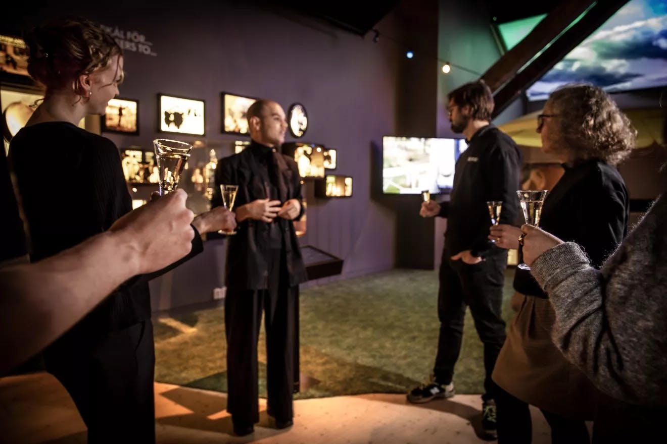 Un groupe de personnes dans une pièce faiblement éclairée, avec des images encadrées au mur, tenant des verres et engageant la conversation.