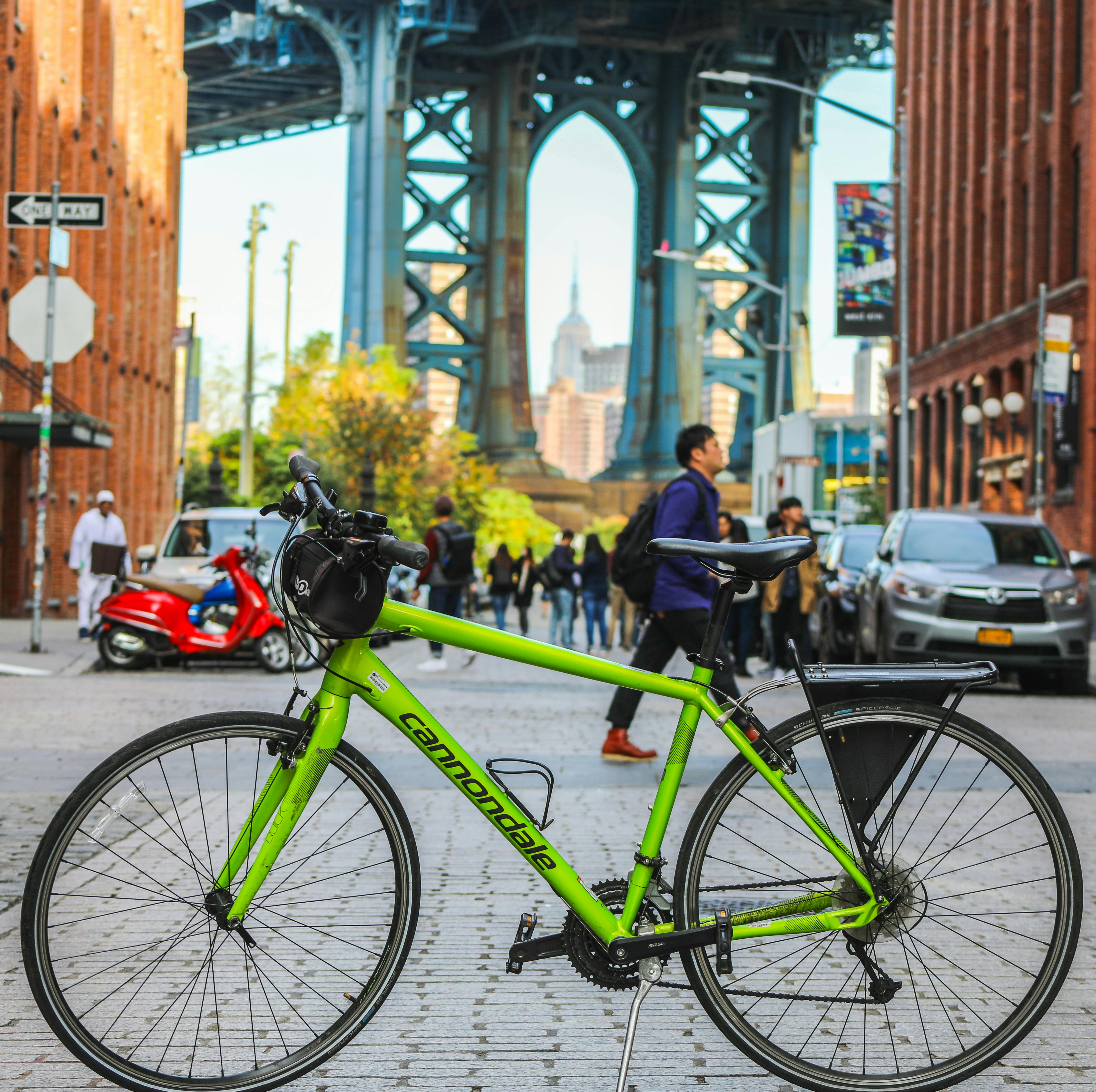 Una bicicletta verde è parcheggiata in primo piano in una scena di strada sotto un grande ponte, con pedoni e automobili sullo sfondo.