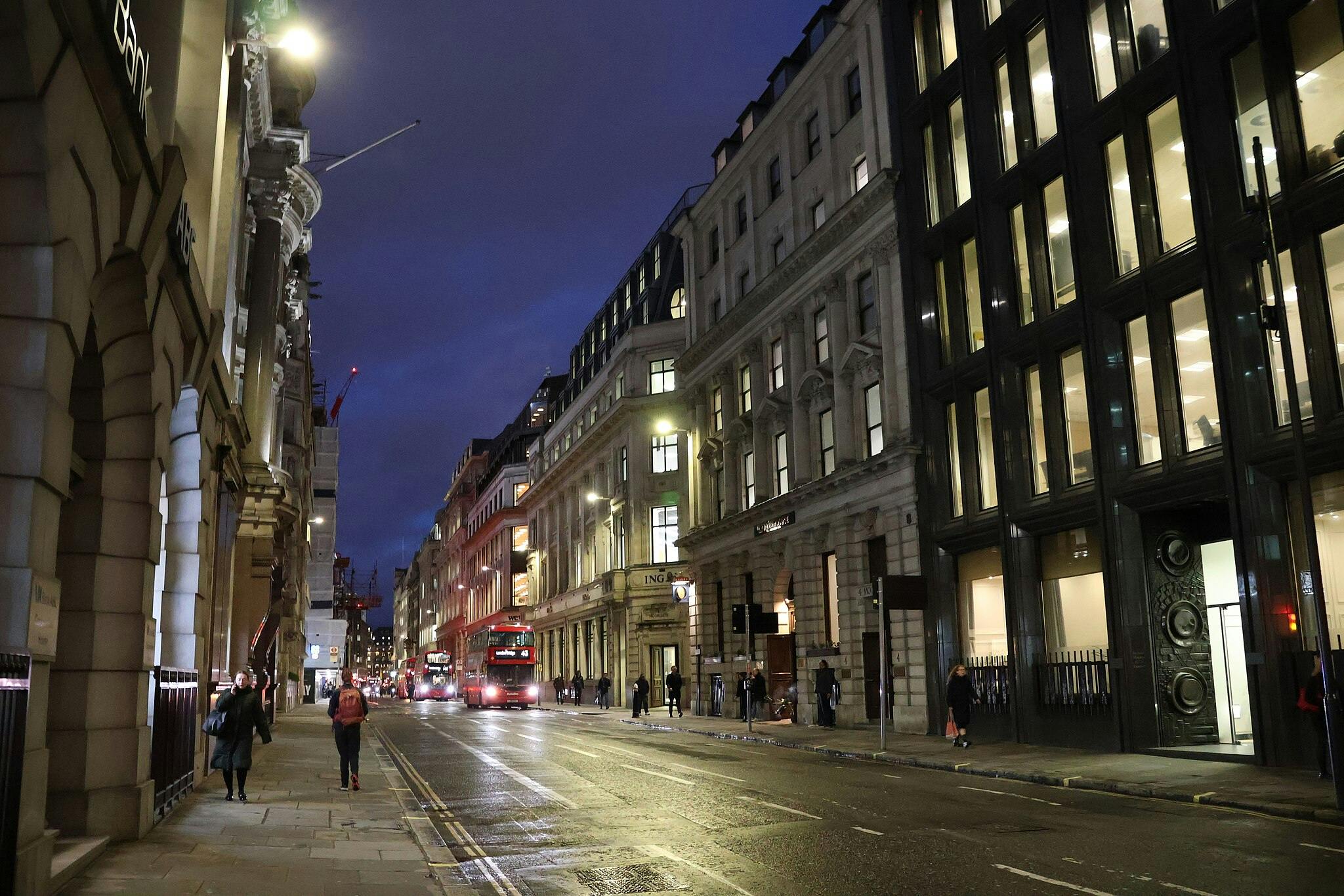 City street at dusk with people walking, illuminated buildings, and double-decker buses in the distance.