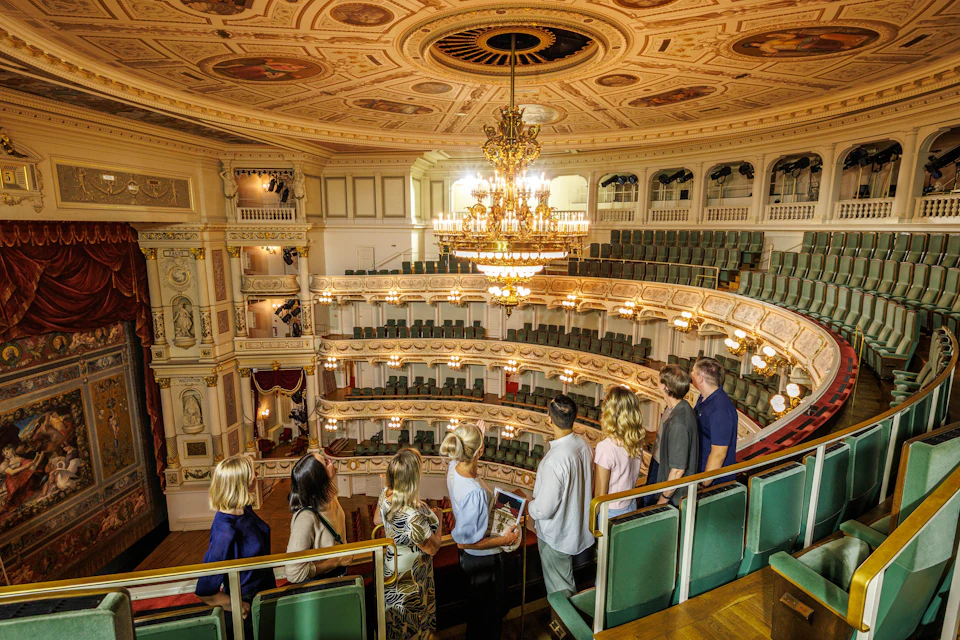 A group of people touring an ornate theater with a grand chandelier and tiered seating under a decorative ceiling.