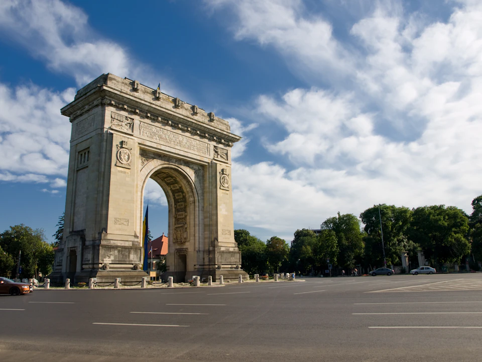 Stone arch monument with decorative carvings on an empty roundabout, surrounded by trees under a partly cloudy sky.