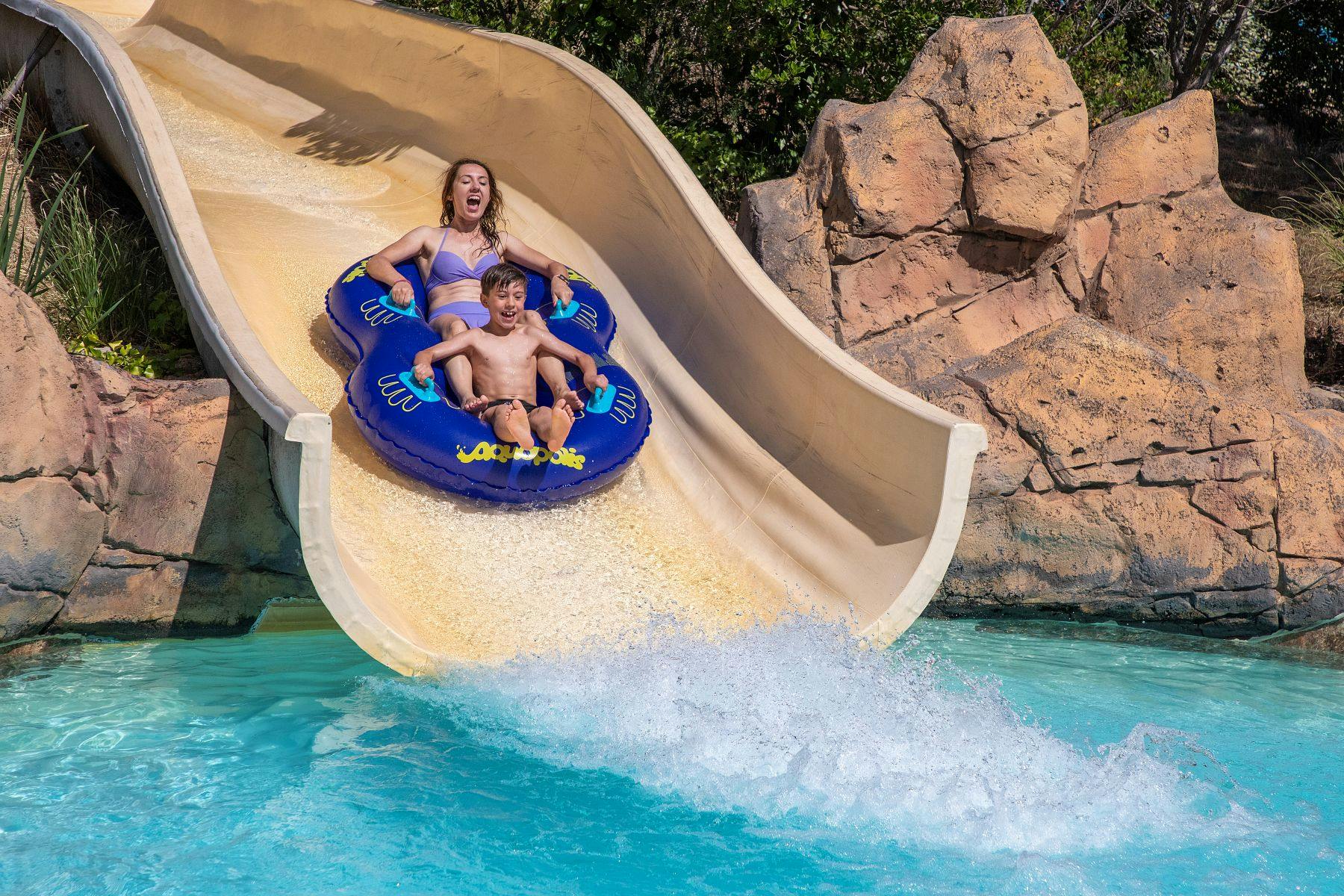 Une femme et un enfant dévalent un toboggan aquatique sur un radeau gonflable bleu, avant de plonger dans la piscine en contrebas.