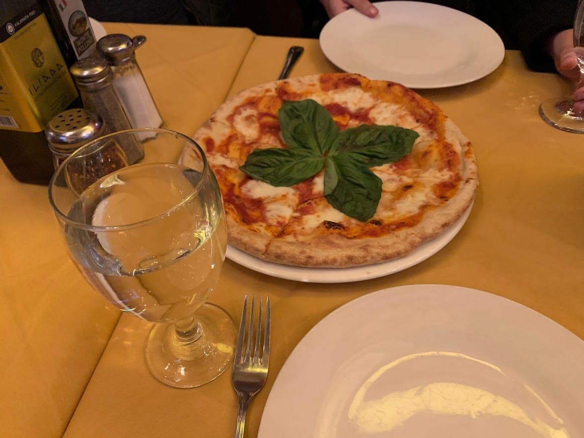Een margherita pizza gegarneerd met verse basilicum op een geel tafelkleed, vergezeld van een glas water en twee lege borden.