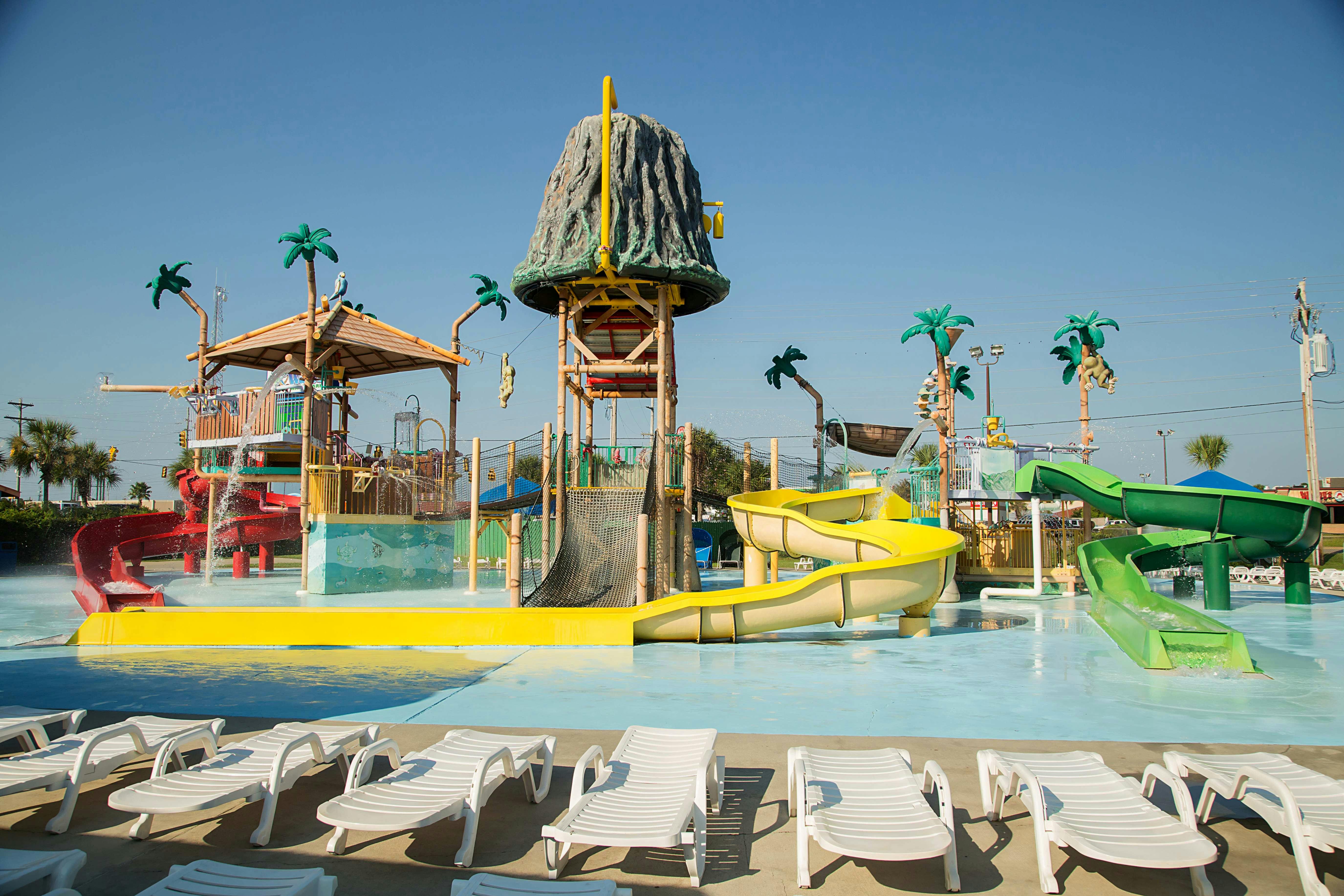 Water park with a volcano-themed play structure, yellow slides, and palm trees, surrounded by empty lounge chairs.