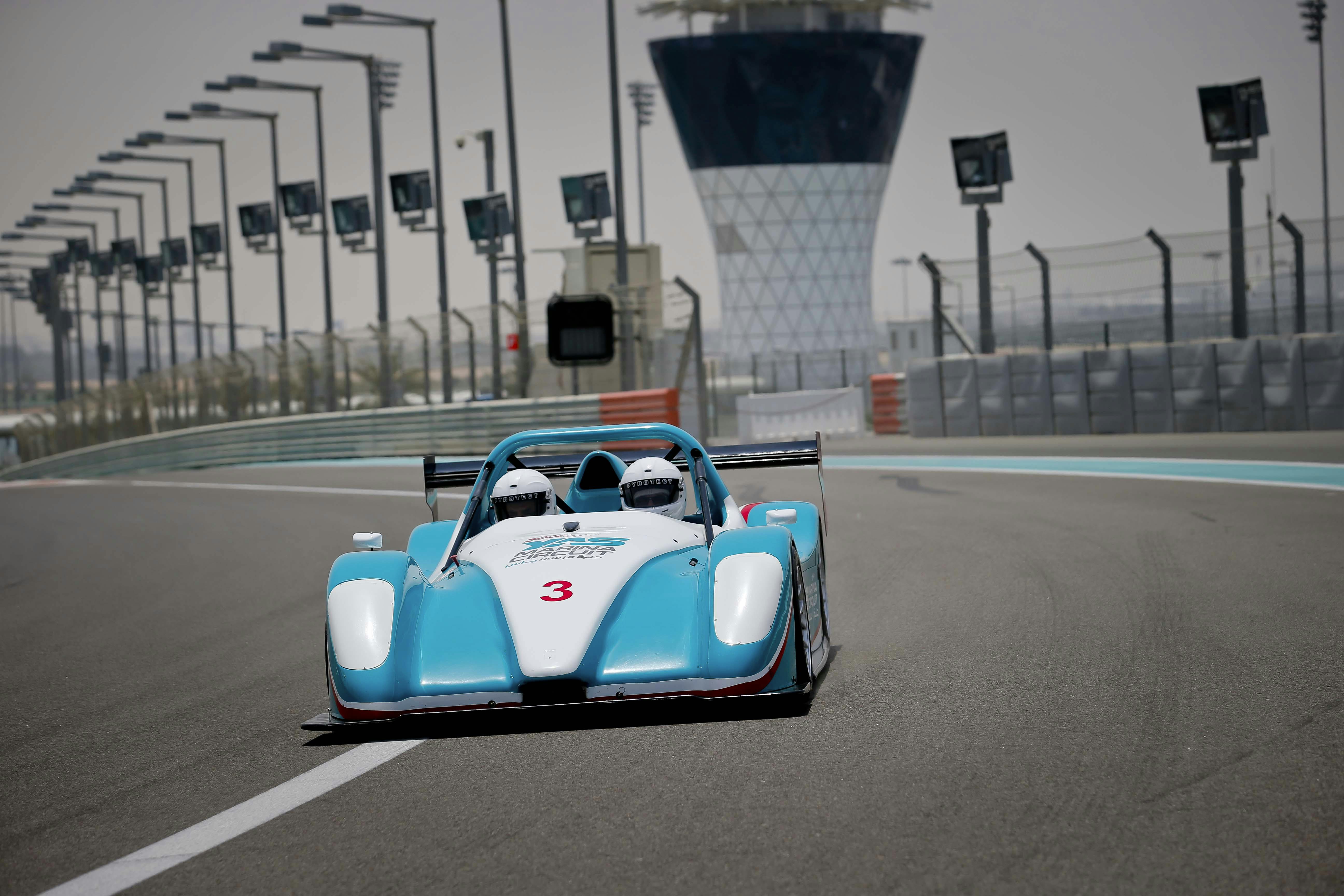 Two helmeted drivers in a blue and white race car on a track with a tower and lights in the background.