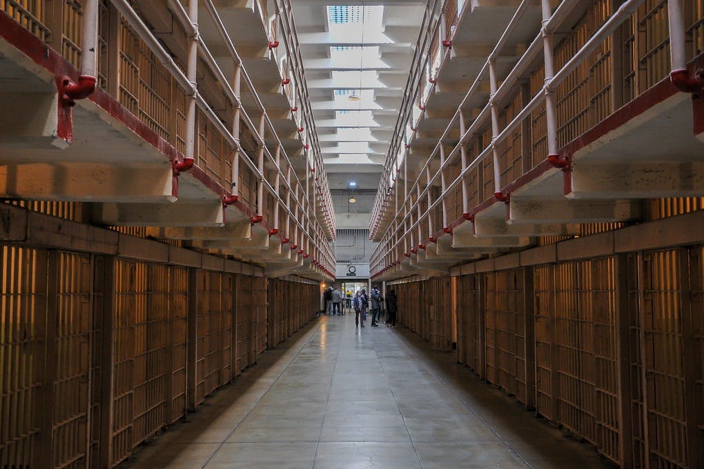 En lång, tom fängelsekorridor med rader av celler på båda sidor och några personer samlade längst bort.