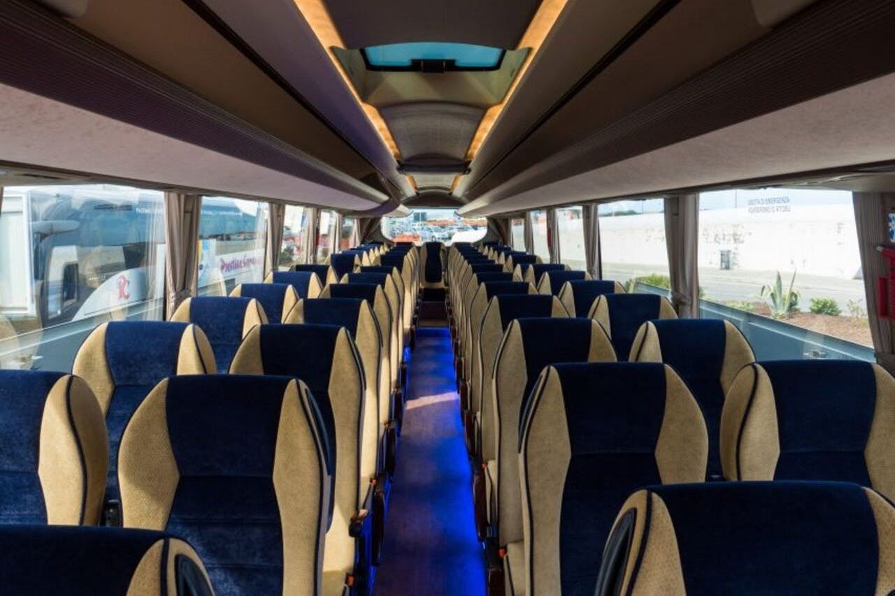 Interiér moderního autobusu s řadami modrých a béžových sedadel, osvětlením uličky a velkými okny na obou stranách.