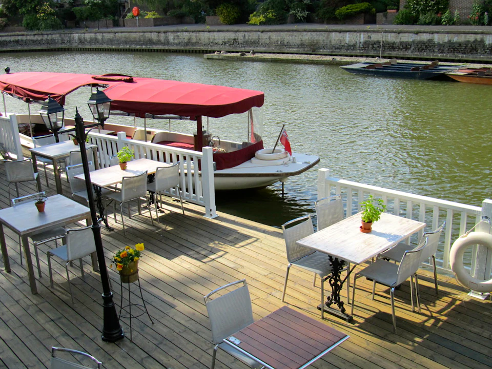 Outdoor wooden deck with chairs and tables overlooking a canal with boats, including one shaded by red canopies.