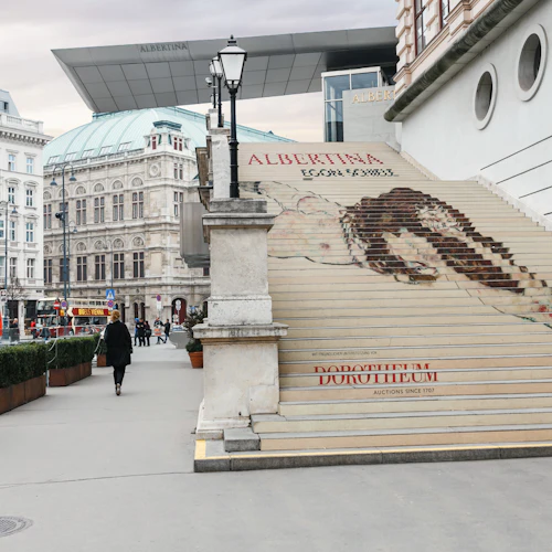 En person går förbi Albertina-museets trappor med ett stort konstverk utställt, medan utsmyckade byggnader kantar bakgrunden.