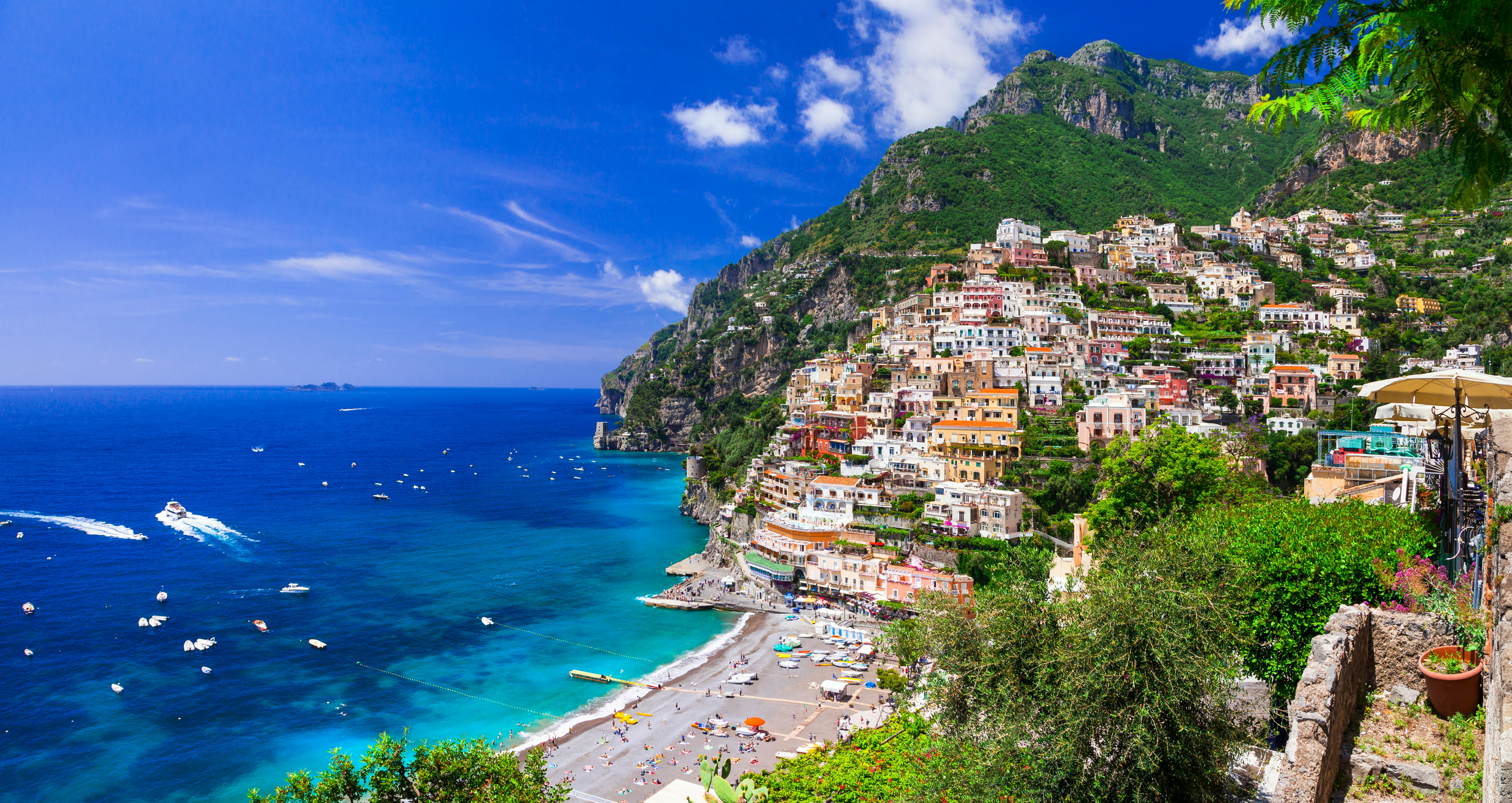 Colorful hillside town overlooking a vibrant blue sea with numerous boats; beach with umbrellas and people in foreground.