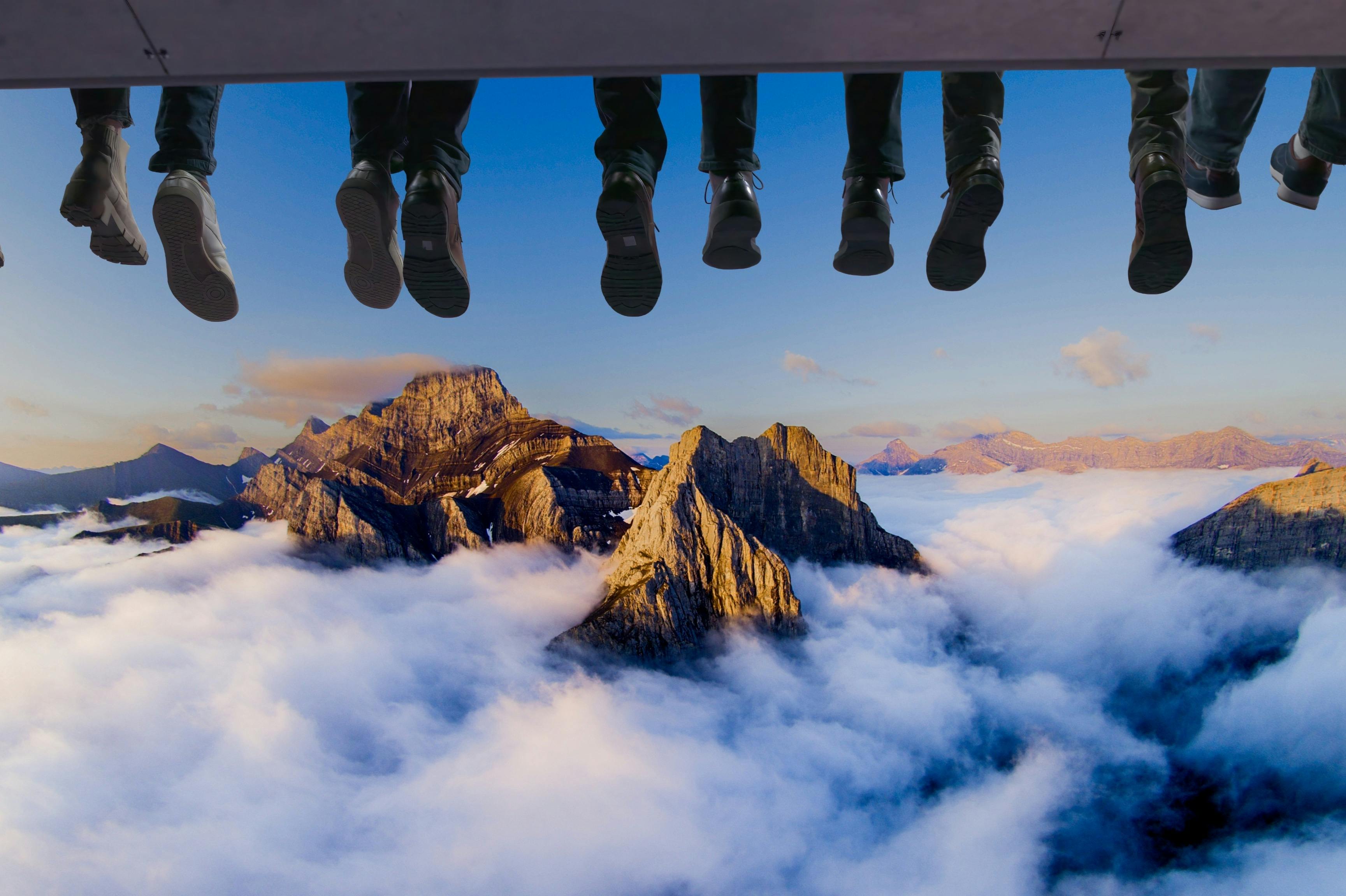 Feet dangling from above against a backdrop of mountains emerging from clouds under a blue sky.