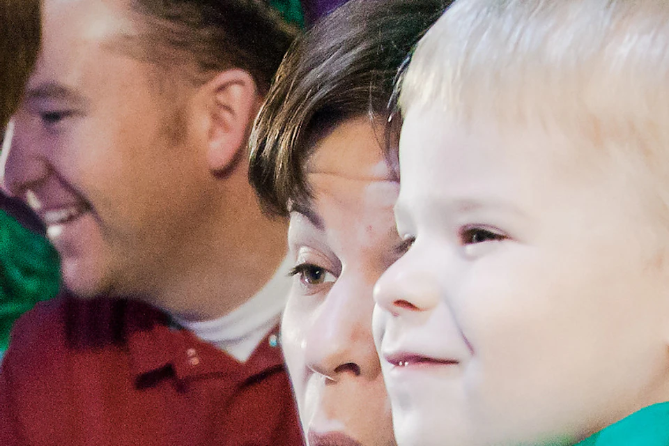 Child pointing at a stingray in an aquarium tank, with an adult and another child closely observing. Purple background.