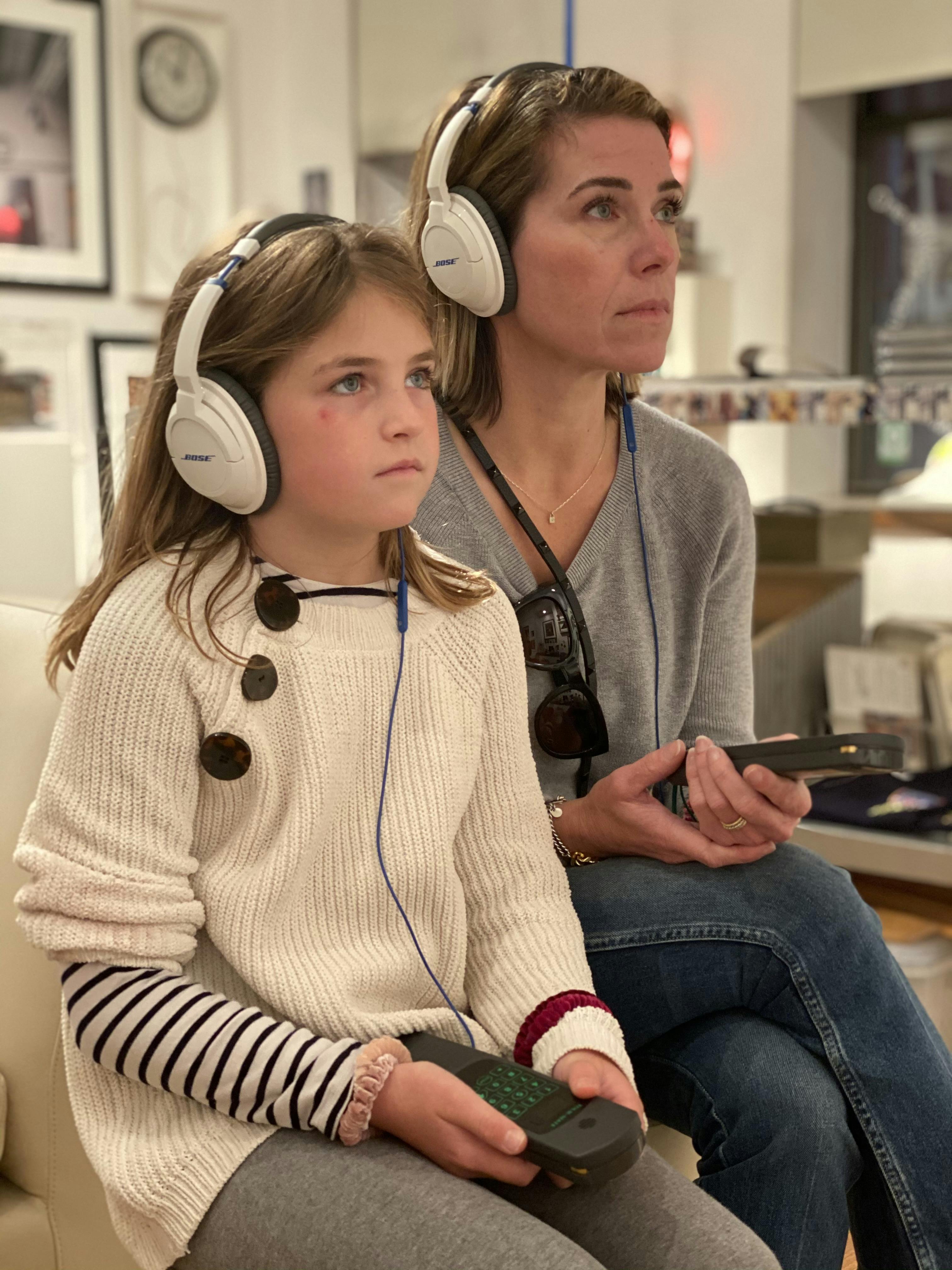 A woman and a girl both wearing headphones sit together, focusing intently. The woman holds a device. Indoor setting.