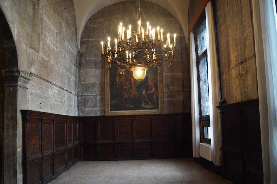 A dimly lit, antique room with stone walls, wooden wainscoting, a large chandelier, and a framed painting above a carpeted floor.