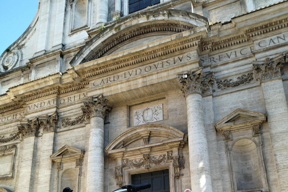 Ornate facade of an ancient stone building with large columns, detailed carvings, and Latin inscription above the doorway.