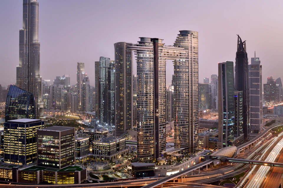 Cluster of modern skyscrapers with illuminated windows, including two connected towers; dusk sky in the background.