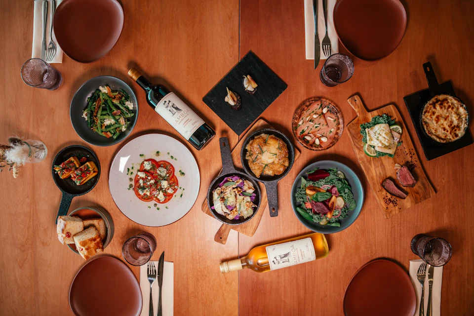 A variety of dishes and drinks arranged on a wooden table, including salads, appetizers, and two bottles of wine.