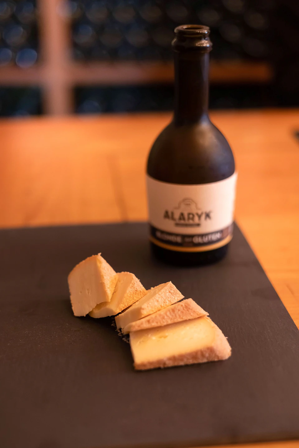 Slices of cheese on a black surface with a bottle labeled "Alaryk" in the background on a wooden table.