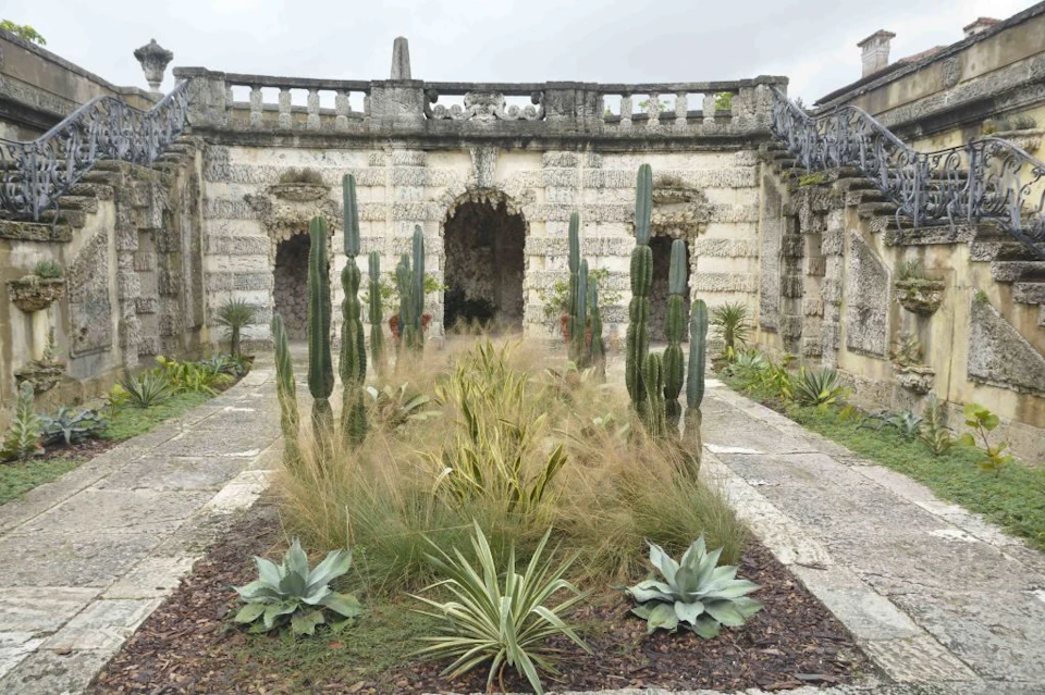 Stone courtyard featuring tall cacti, ornamental grasses, and a weathered stone building with arched doorways and railings.