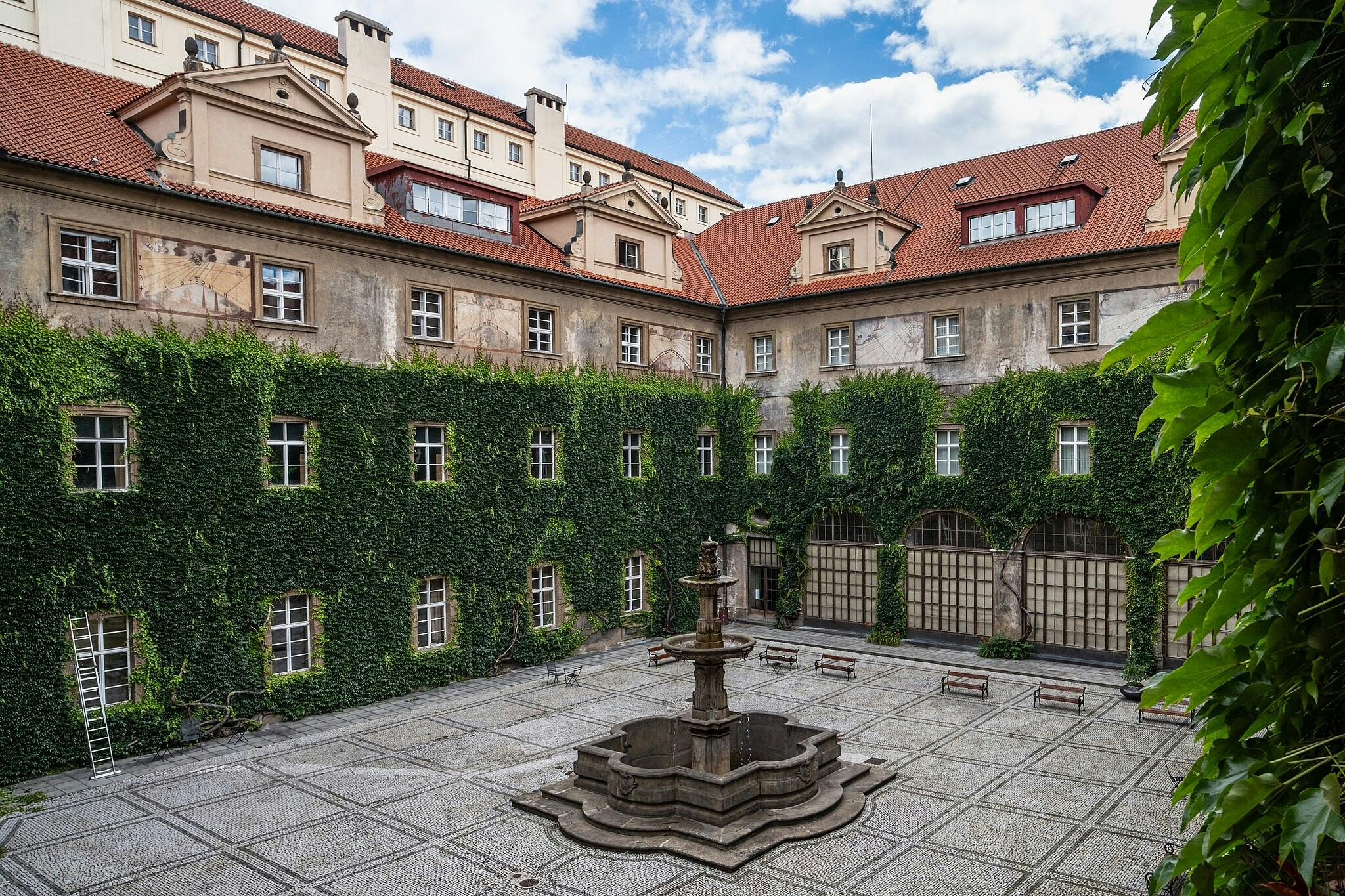 Patio con muros cubiertos de hiedra, fuente central de piedra, bancos, escalera y edificio antiguo con tejado y ventanas de tejas rojas.
