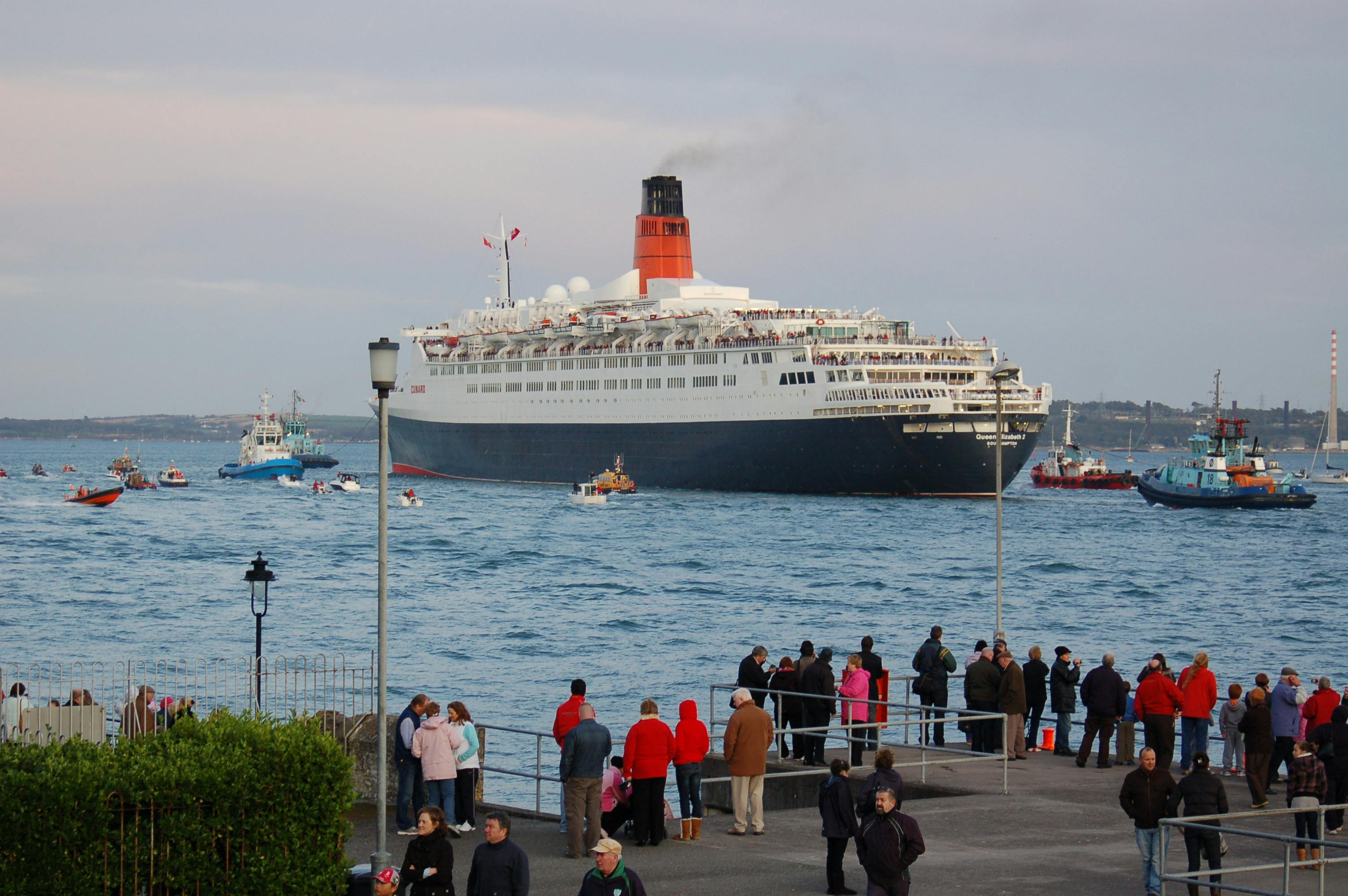 A large cruise ship with a red funnel passing by a waterfront, with people standing and watching from the shore.