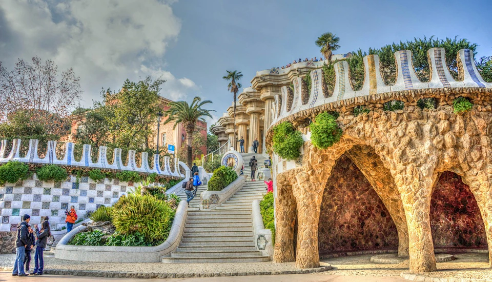 People walking on grand, ornate stairs surrounded by lush greenery and unique stone structures with a cloudy sky above.
