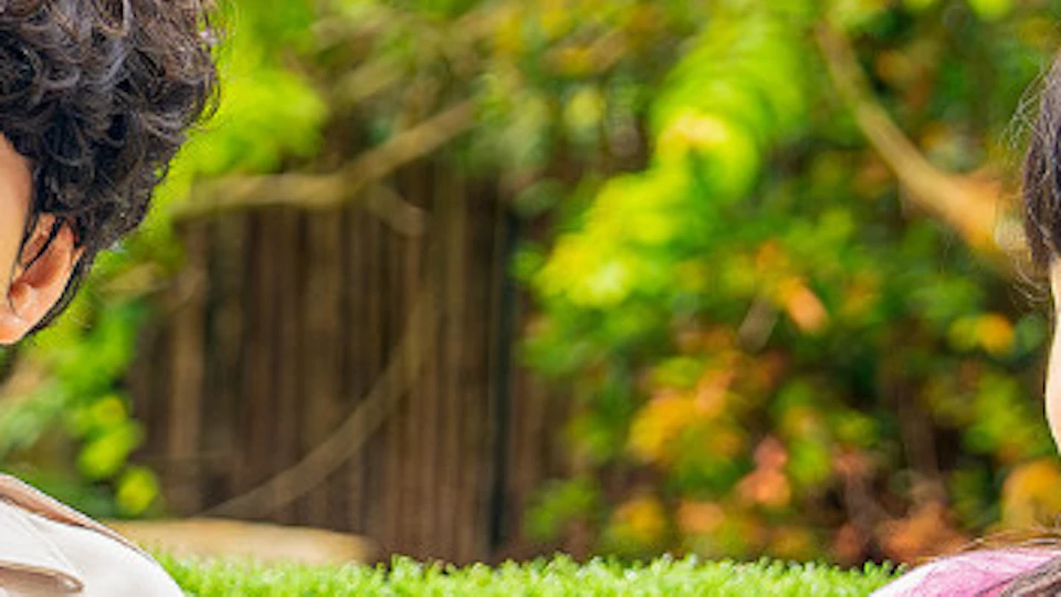 Two children stand outdoors beside a green hedge, pointing and smiling, with trees and a wooden fence in the background.
