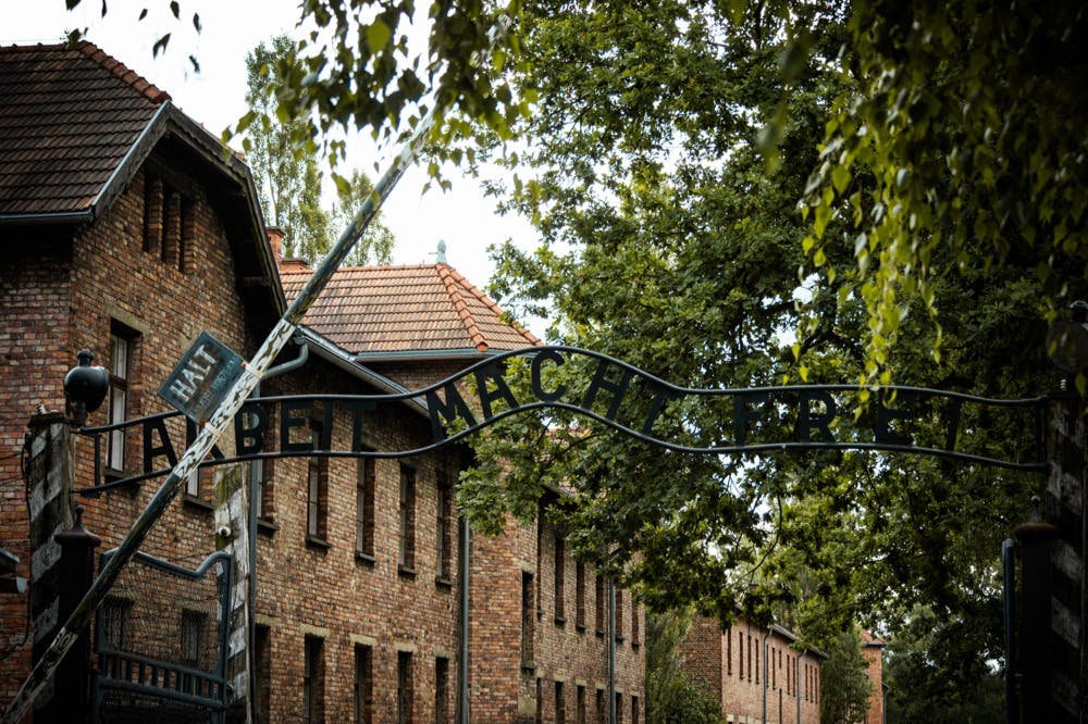 小径上有铁门，上面刻有“Arbeit Macht Frei”字样，周围环绕着红砖建筑和郁郁葱葱的绿树。