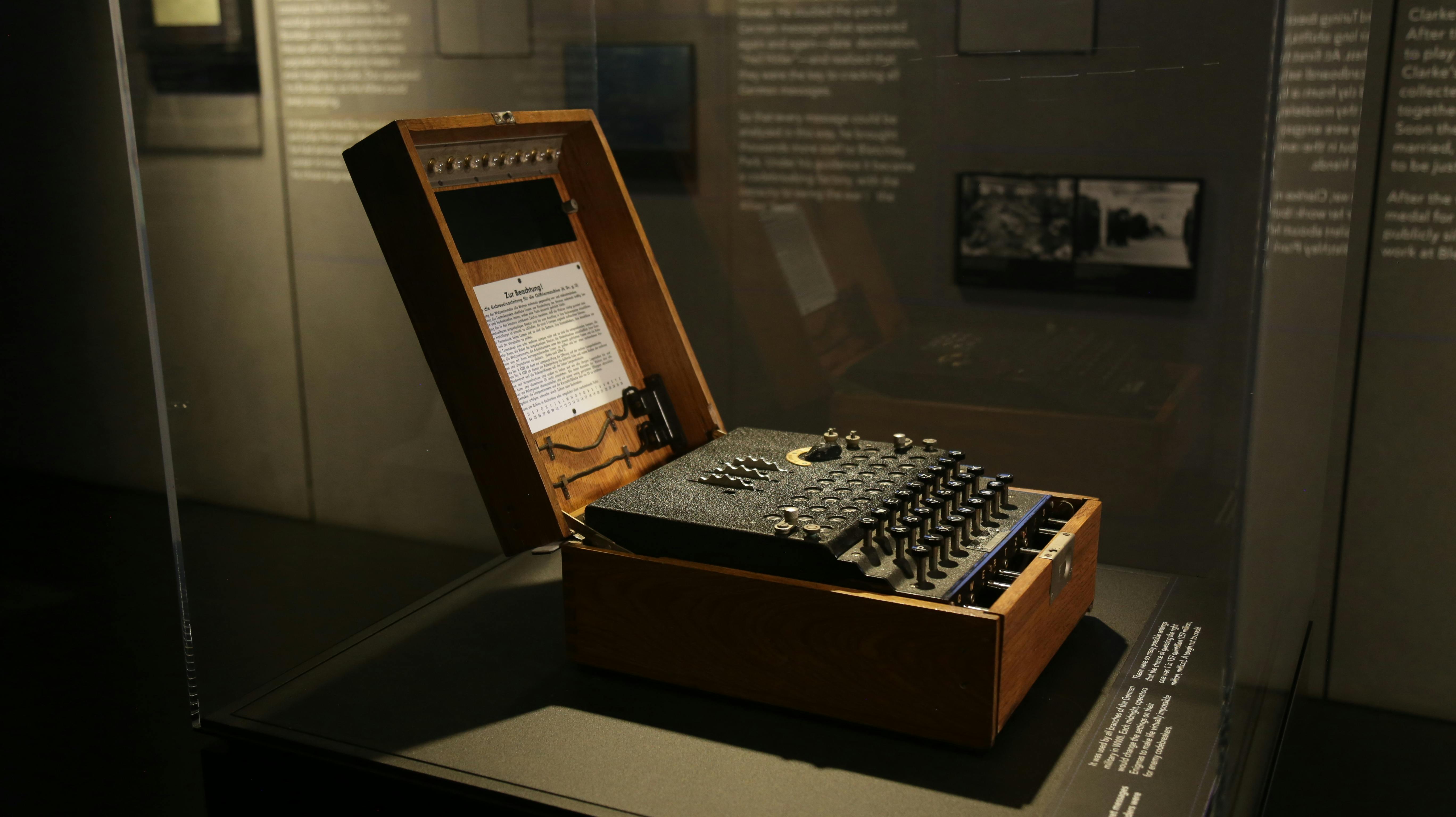 Ένα μηχάνημα Enigma σε ξύλινη θήκη εκτίθεται σε ένα μουσειακό έκθεμα, με κείμενο πληροφοριών ορατό μέσα στο καπάκι.