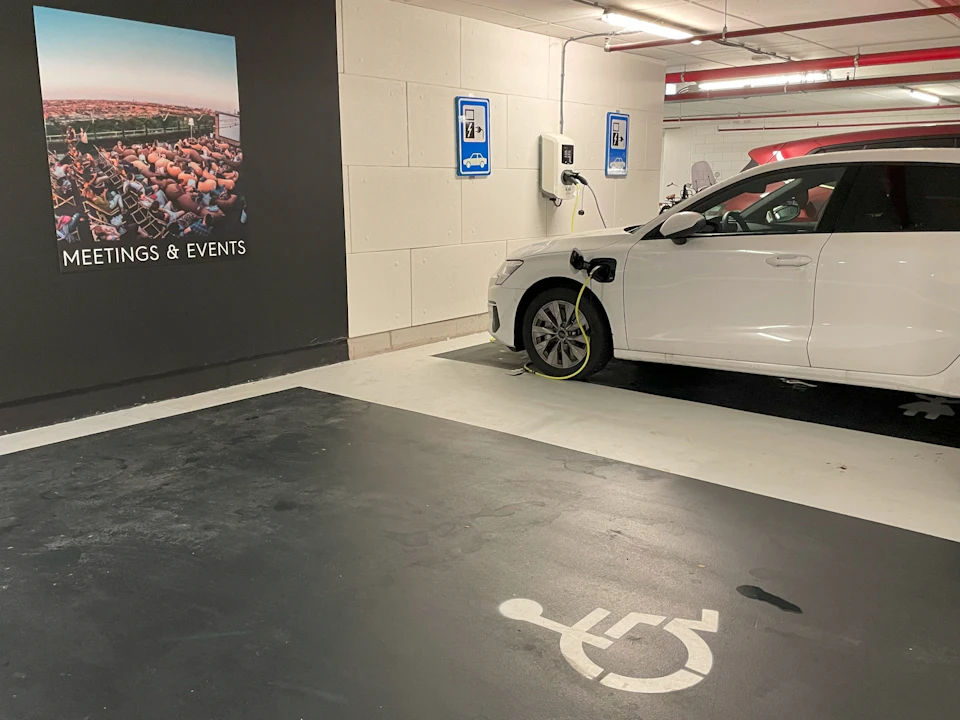 A white car is charging at an electric vehicle station in a parking garage. A nearby space is marked with a wheelchair symbol.