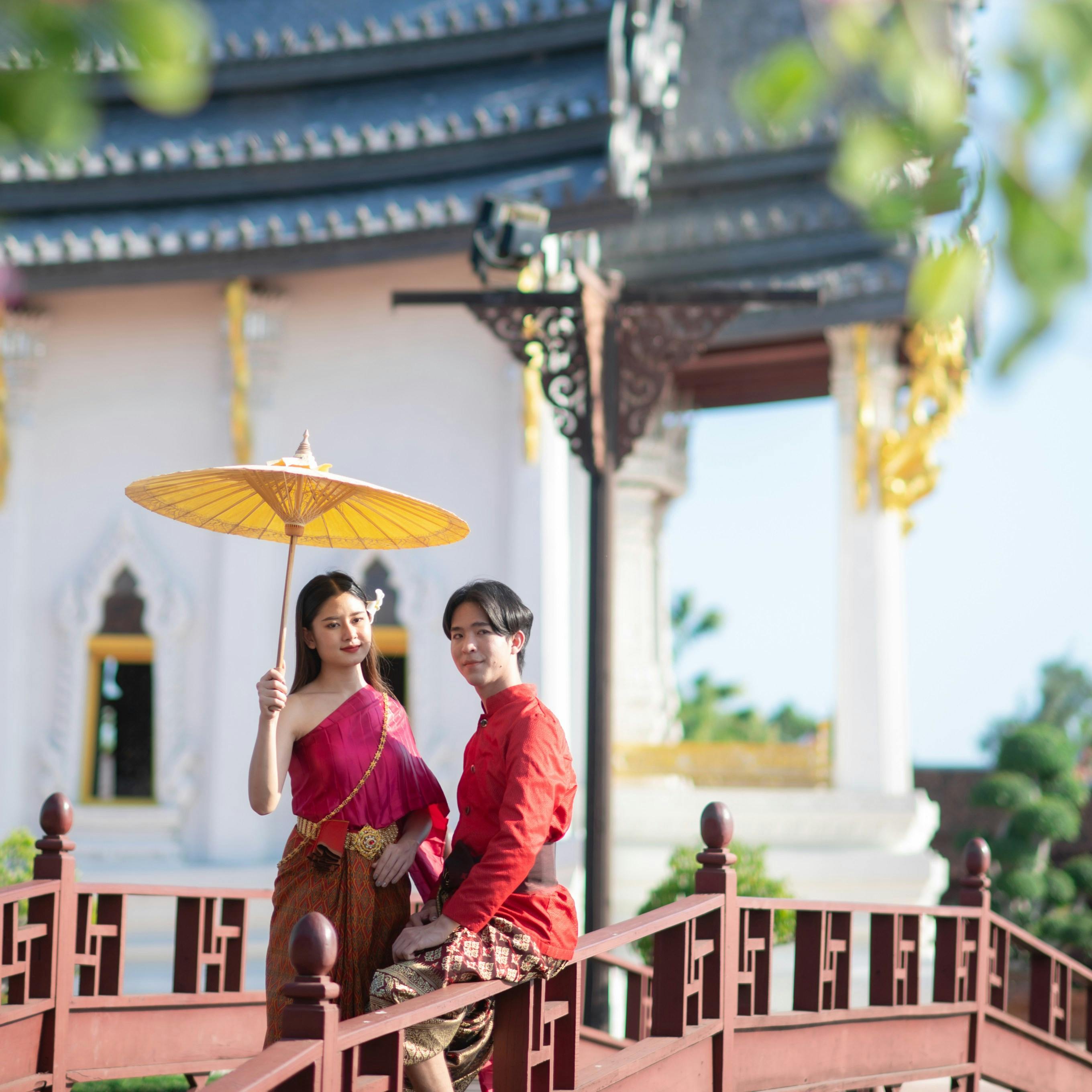 Two people in traditional attire stand on a decorative bridge, one holding a yellow parasol, with a temple in the background.