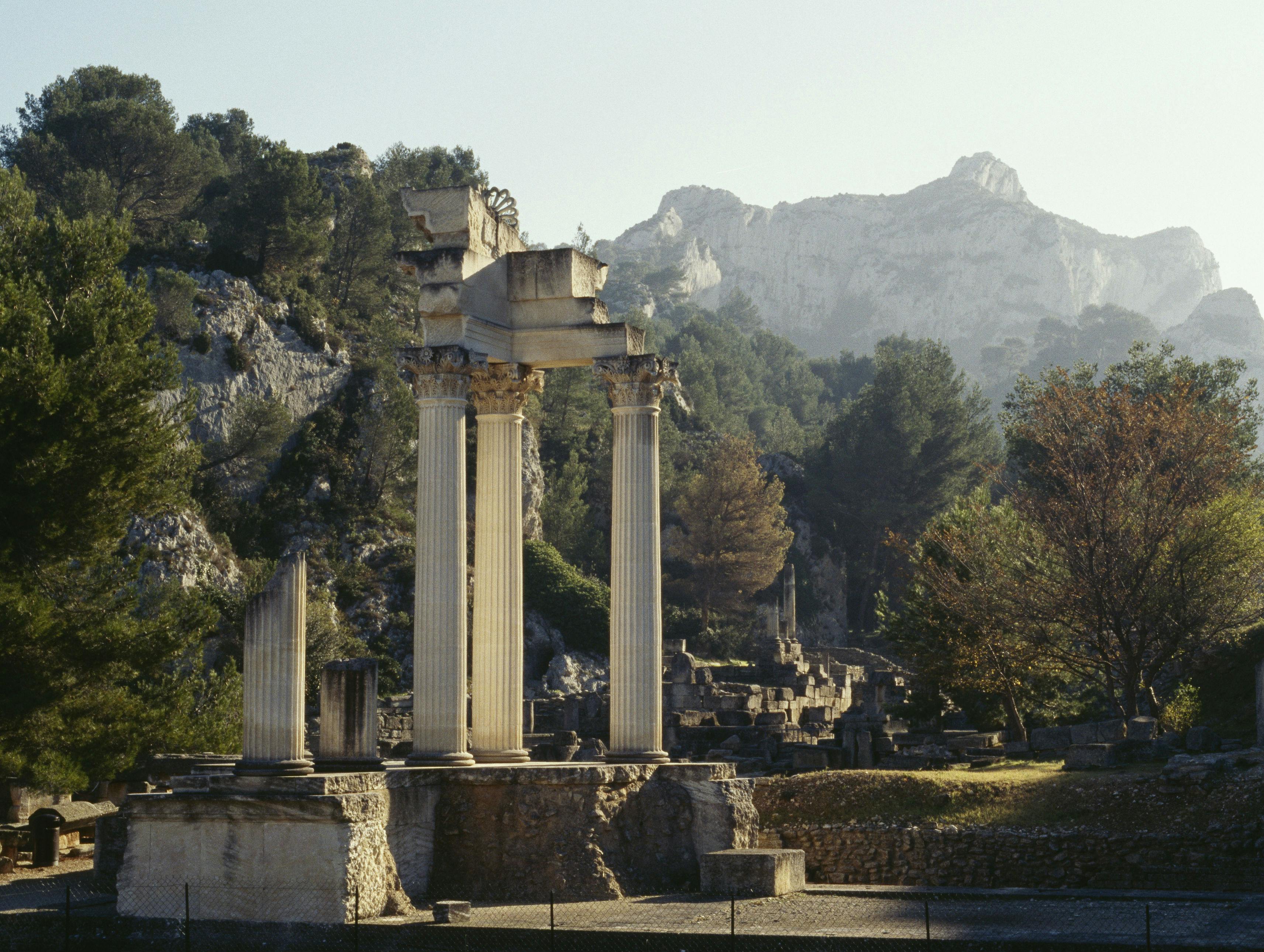 Rovine di antiche colonne e strutture circondate da alberi e montagne sullo sfondo, sotto un cielo limpido.