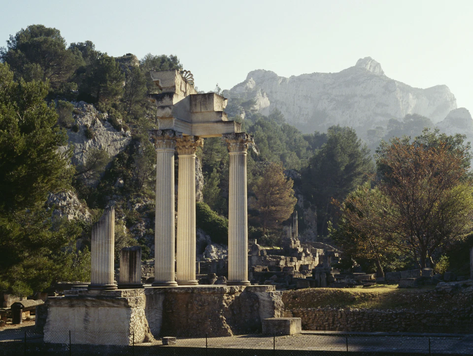 Ruins of ancient columns and structures surrounded by trees and mountains in the background under a clear sky.