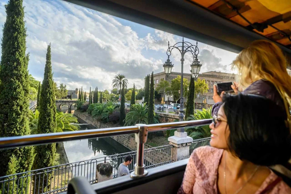 Two women on a bus, one looking out the window and the other taking a photo. Outside, an urban park and old building are visible.