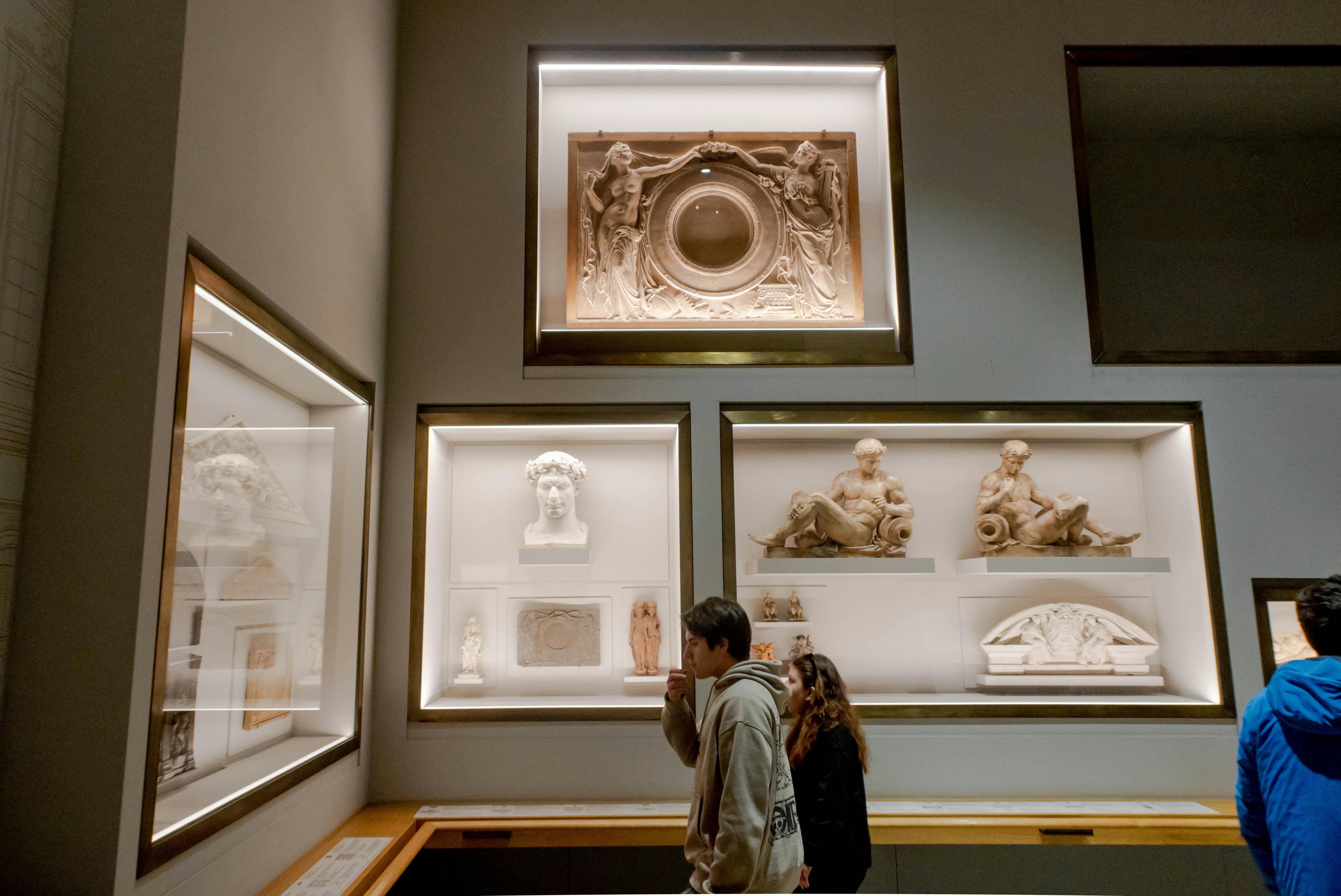 Deux personnes regardent des sculptures classiques et des reliefs exposés dans une galerie de musée bien éclairée.