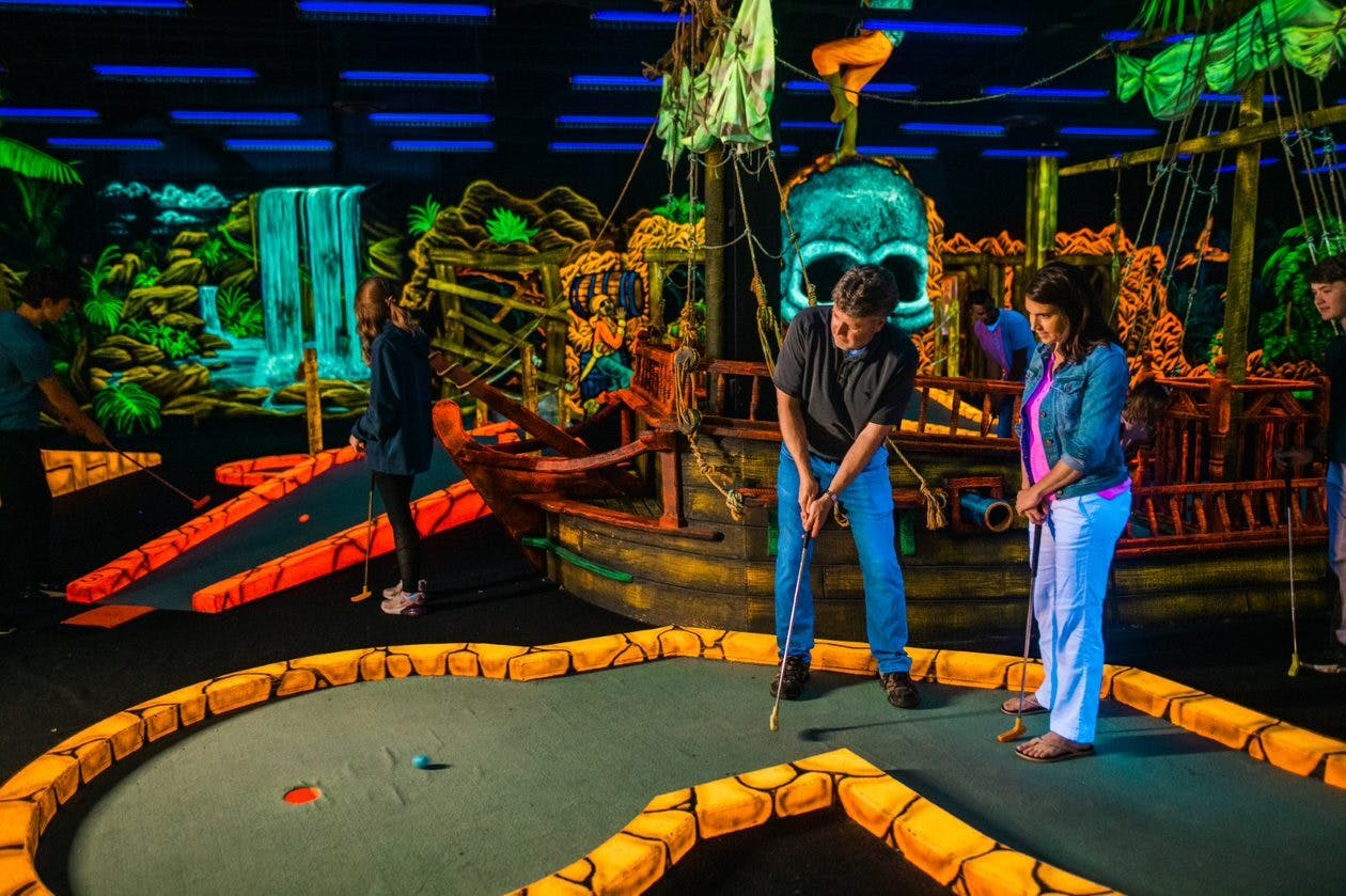Two people playing mini-golf in a pirate-themed indoor course with glowing decorations and a large skull structure in the background.