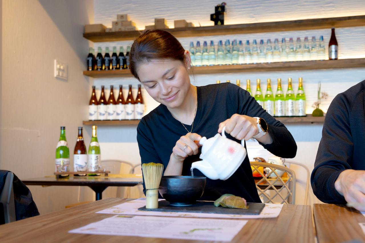 A woman pours liquid from a white teapot into a black bowl on a wooden table, with various bottled drinks on shelves behind her.
