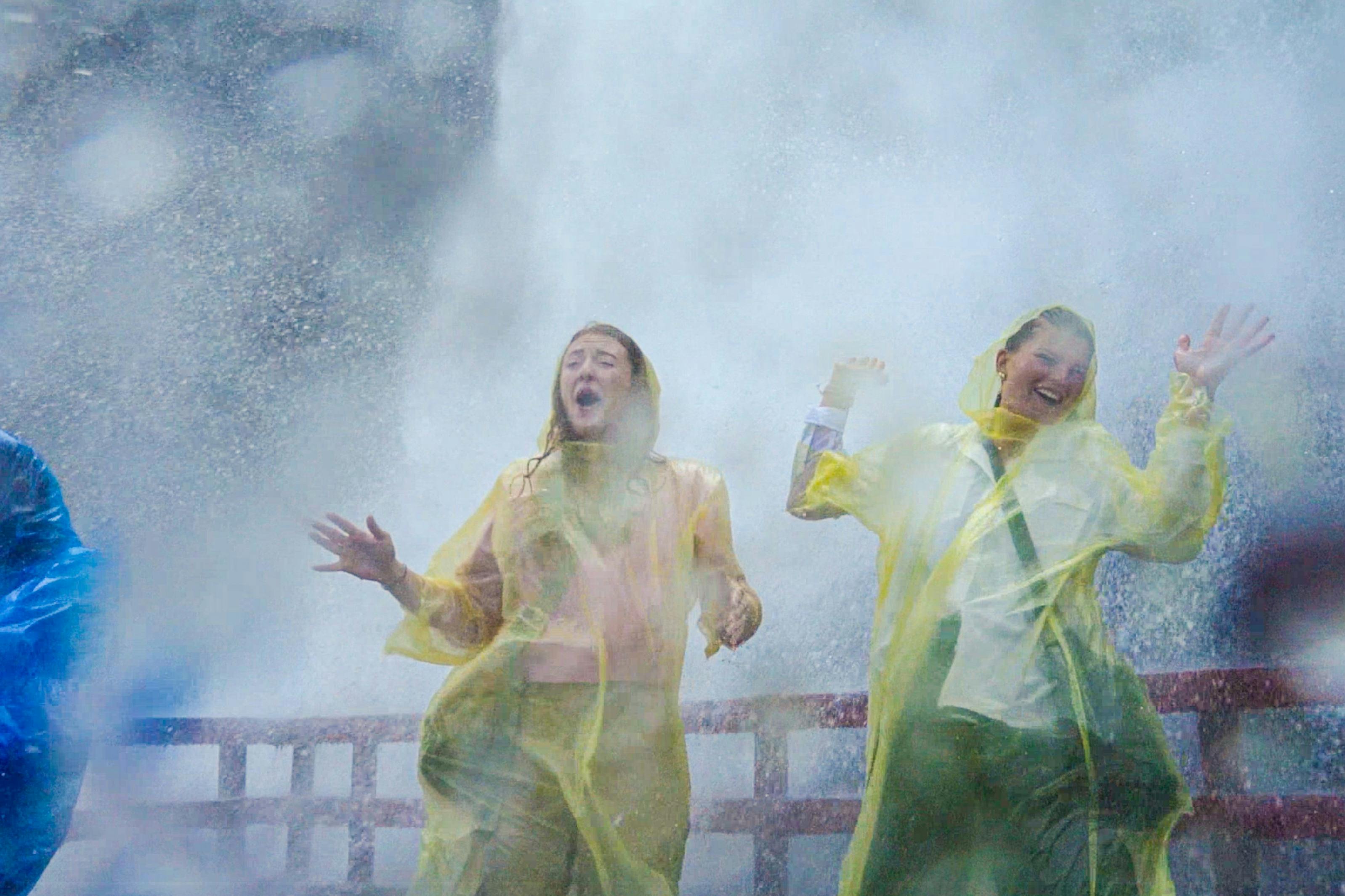 黄色いポンチョを着た2人の人が木製のプラットフォームの上に立ち、大きな水しぶきが当たると喜んで反応します。