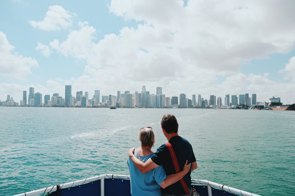 Two people standing on a boat, embracing while looking at a city skyline across the water under a partly cloudy sky.