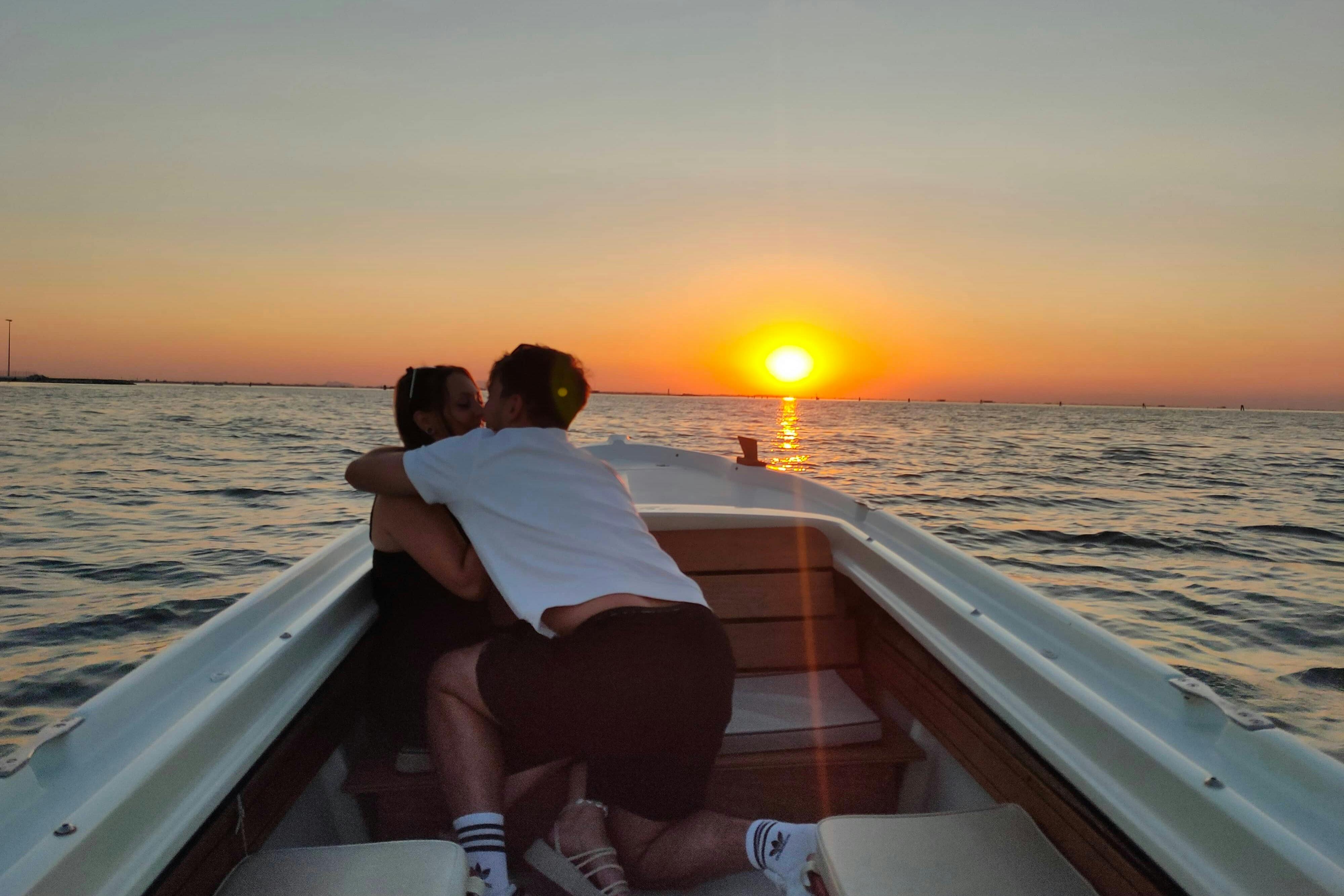 Twee mensen omhelzen elkaar op een kleine boot tijdens een zonsondergang boven kalm water.