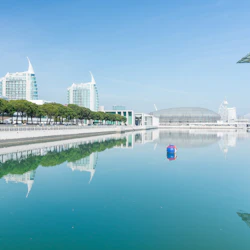 En lugn vattenväg återspeglar moderna byggnader, träd och en blå boj under en klarblå himmel.