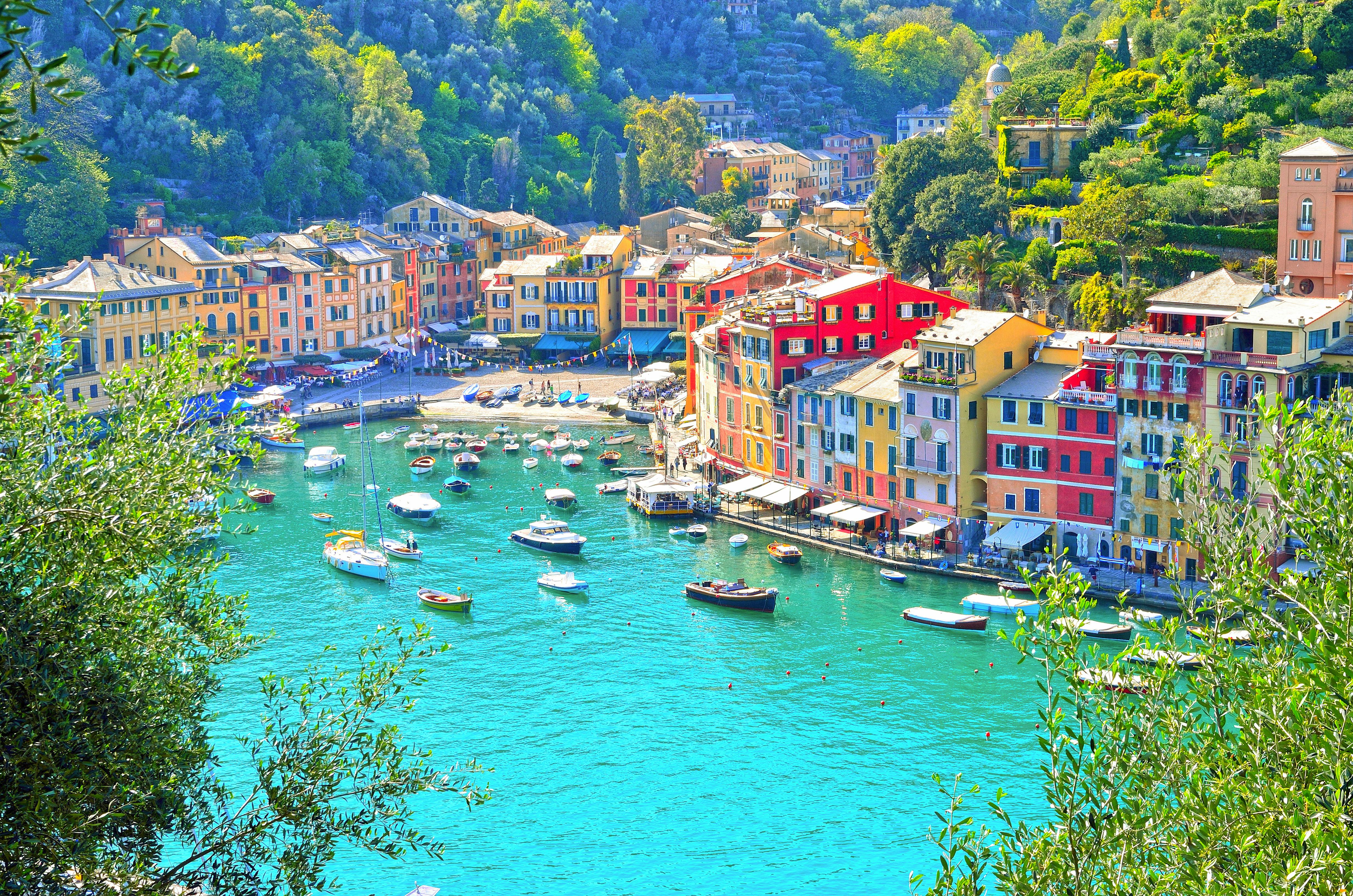 Colorful buildings line a harbor with numerous boats on vivid blue water, surrounded by lush green hills.