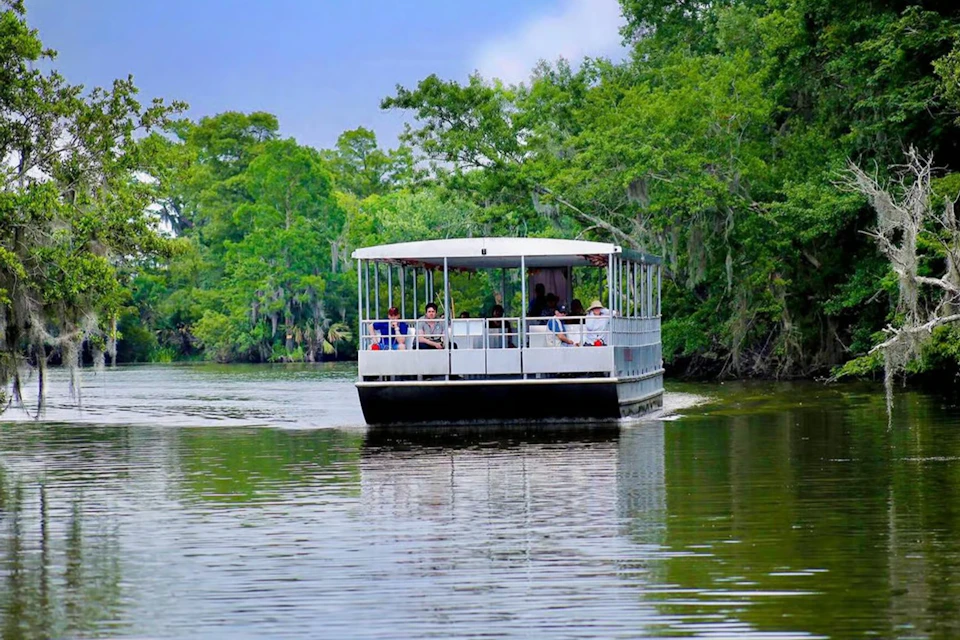 A flat-bottomed tour boat with passengers cruises on a calm river surrounded by lush green trees under a cloudy sky.