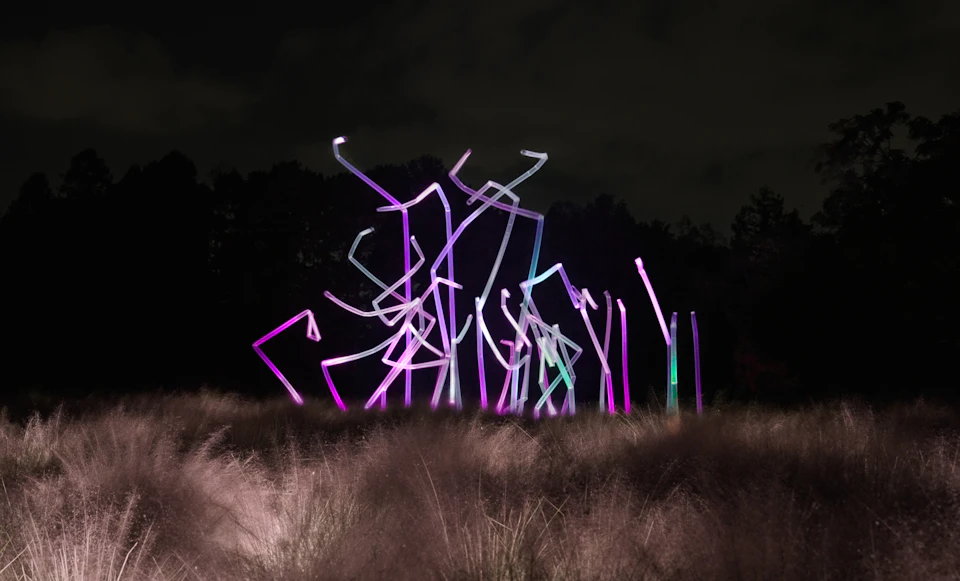 Illuminated abstract sculpture with multicolored lights in a grassy field at night, trees in the background.